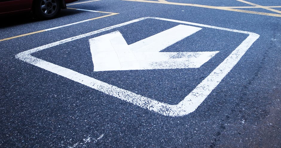 A close-up view of a designated parking bay on an asphalt surface, featuring a large white arrow painted within a square outline, indicating the direction for vehicles to proceed. The painted markings